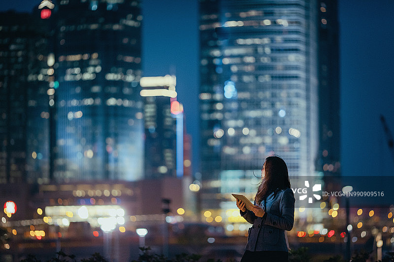 年輕的女商人在夜晚使用數碼平板電腦，自信地回顧著燈火通明的城市天際線圖片素材