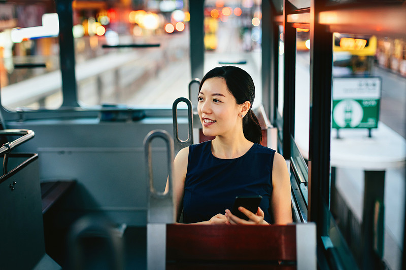 美麗的年輕亞洲女士在城市乘坐公共交通工具時，用智能手機通過窗戶欣賞城市風景圖片素材