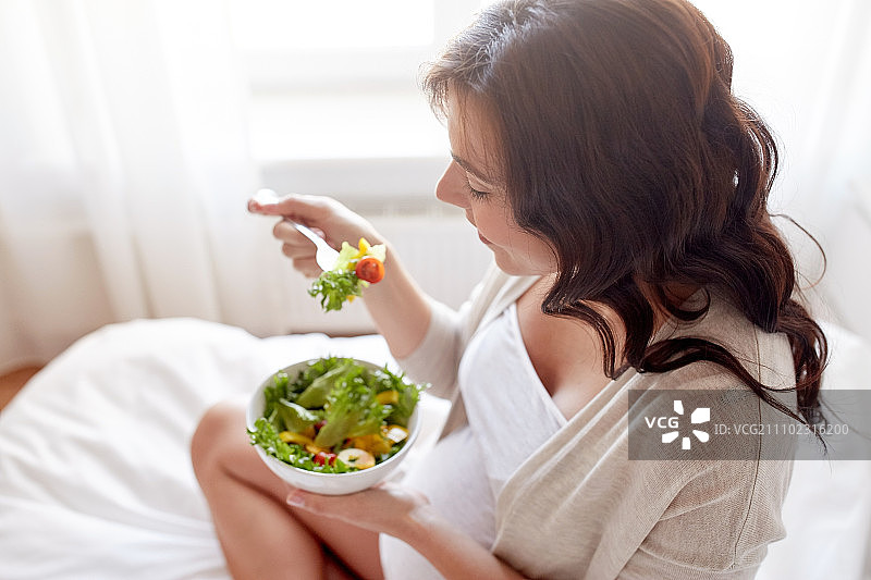 怀孕，健康的食物和人们的概念-近距离观察幸福的孕妇吃蔬菜沙拉早餐在床上在家图片素材