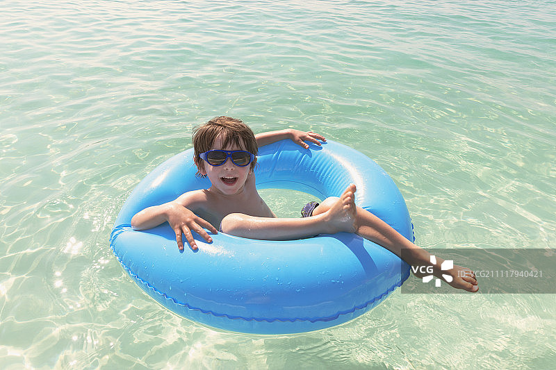 肖像微笑的男孩戴着太阳镜漂浮在蓝色的充气环在海洋图片素材