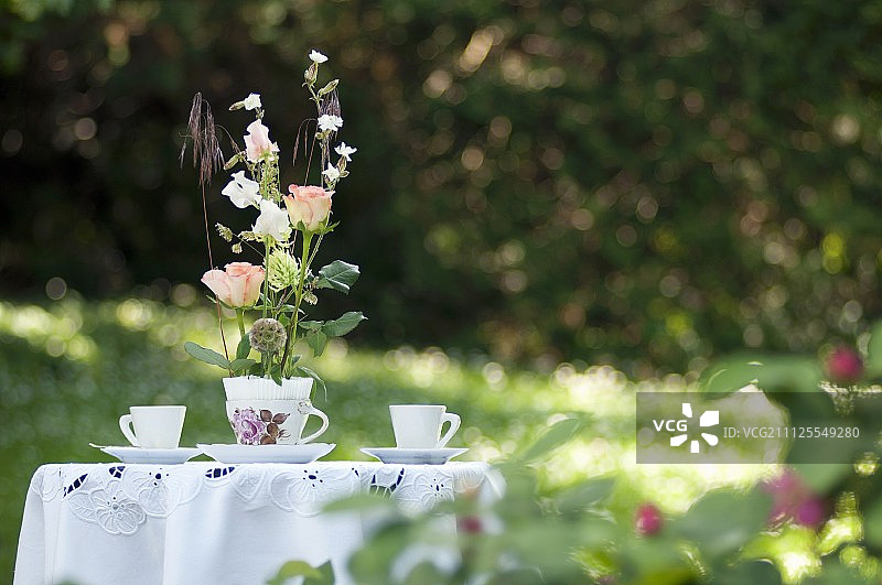 浪漫的玫瑰在怀旧的茶杯安排在白色桌布和两个茶杯的花园桌上图片素材
