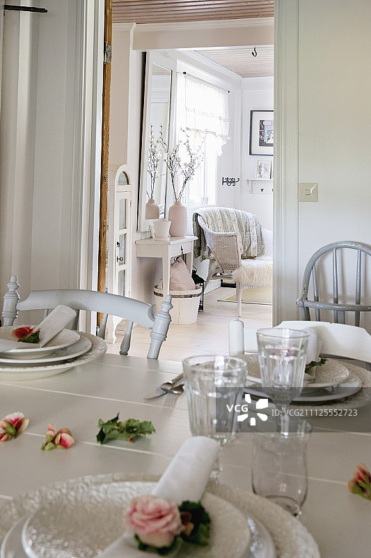 将餐具和玫瑰放在白色的餐桌上;透过背景中打开的门可以看到质朴的客厅图片素材