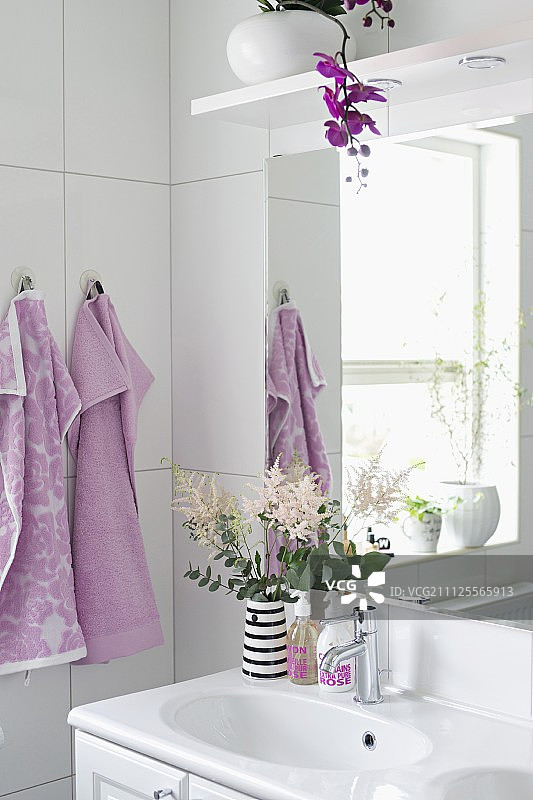水池上放着花瓶，镜子上放着盆兰，白色浴室的挂钩上挂着淡紫色的毛巾图片素材