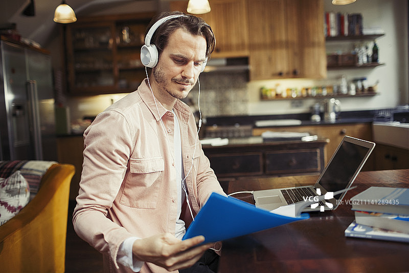 戴着耳机的男人在笔记本电脑前工作，在厨房里阅读文件图片素材
