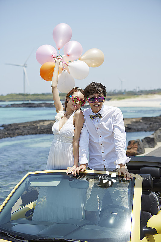 新婚夫妇坐在海边公路上的黄色汽车里，女人拿着气球图片素材