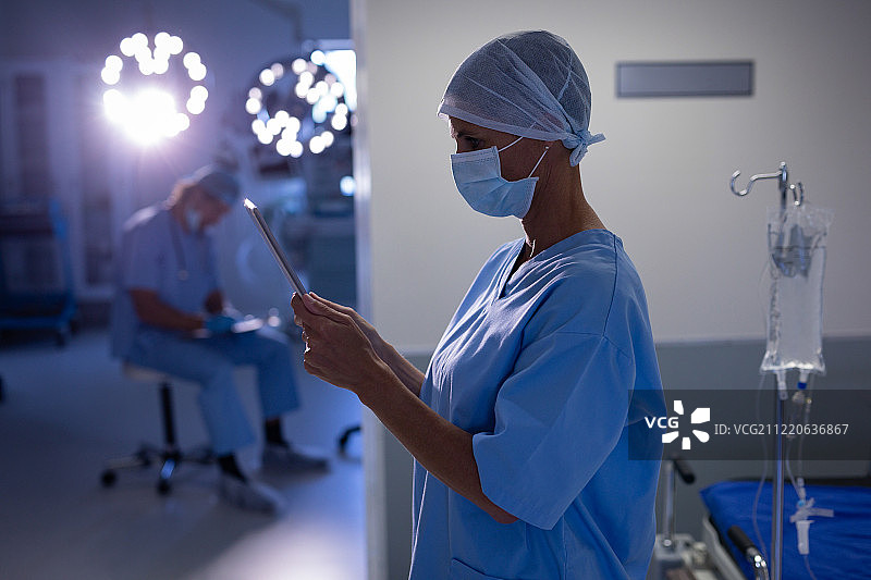 一位白种女外科医生在手术室里使用电子平板电脑的侧视图，而她身后的一名男子正在手术室里写作图片素材