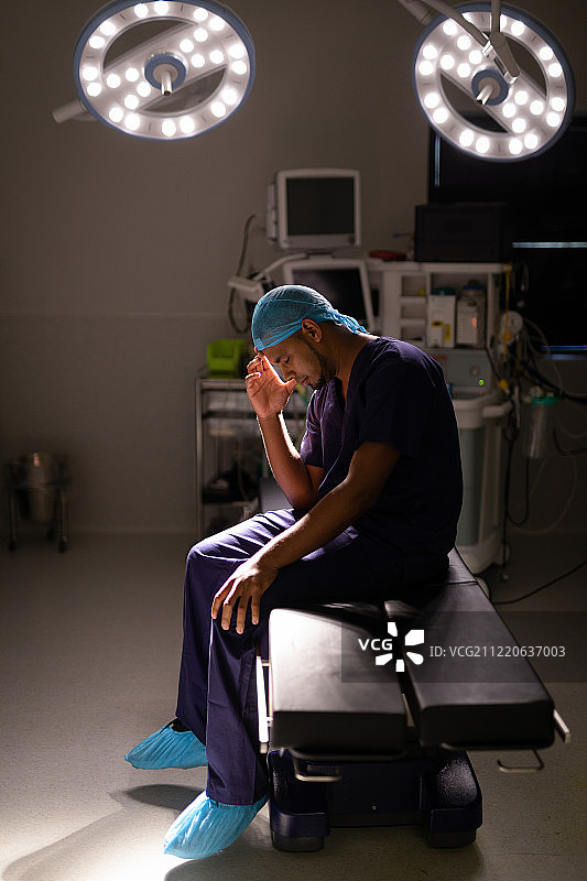一个紧张的非裔美国男性外科医生坐在他的头在他的手在医院的手术室在光线下图片素材