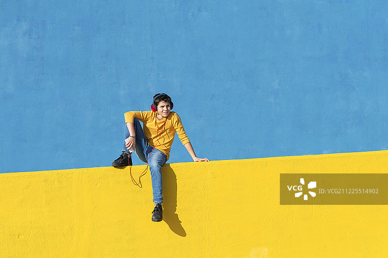 正面的一个小男孩穿着休闲衣服坐在一个黄色的篱笆对着一堵蓝色的墙…图片素材