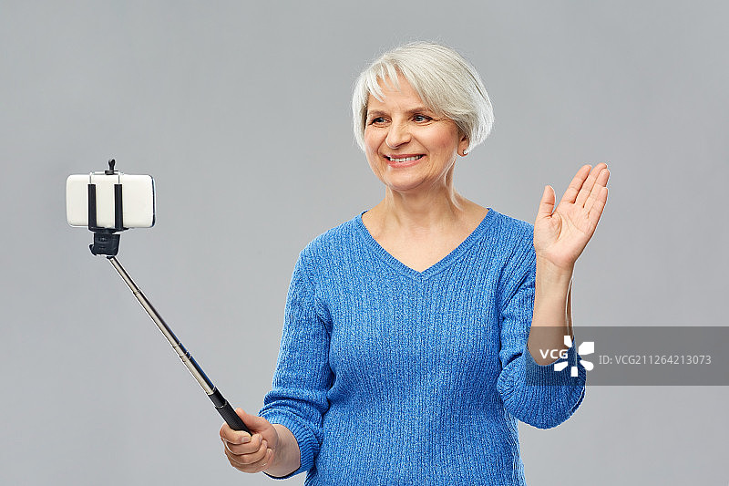 科技与老年人概念——微笑的老年女性用智能手机自拍，在灰色背景上挥动手拍照。微笑的高级女性用智能手机自拍图片素材