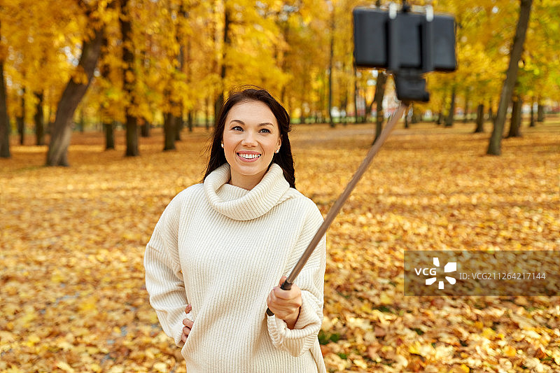 季节，科技和人的概念-美丽的年轻快乐的女人用智能手机拍照在秋季公园的自拍杆。一名女子在秋季公园用智能手机自拍图片素材