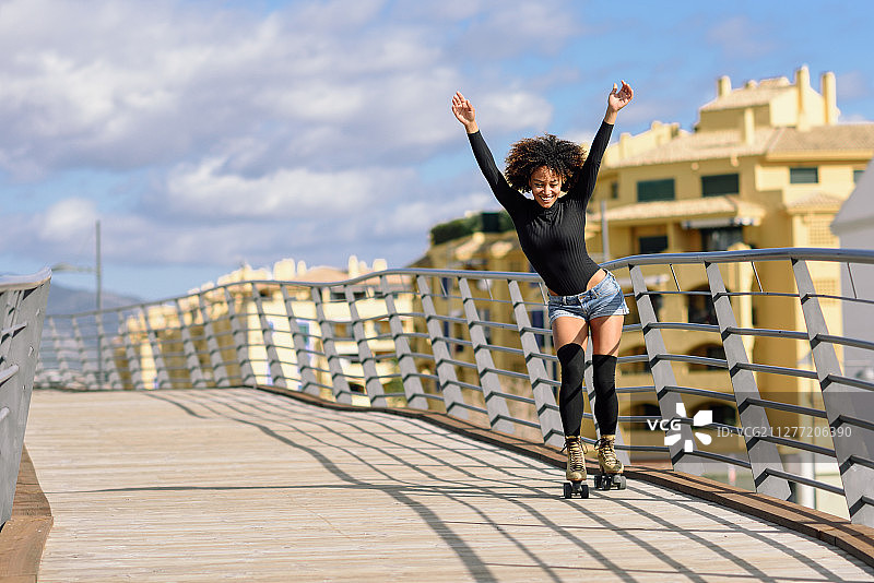 黑人妇女，非洲发式，溜冰鞋在户外城市桥上张开双臂。在阳光明媚的日子里，微笑着滑旱冰的年轻女性。天空中美丽的云彩。非洲发型的女人在户外骑轮滑在城市桥上图片素材