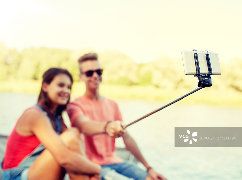 科技，假期和人的概念-快乐的青少年夫妇拍照的智能手机自拍杆和坐在河边的泊位在夏天。一对快乐的青少年情侣在用智能手机自拍图片素材