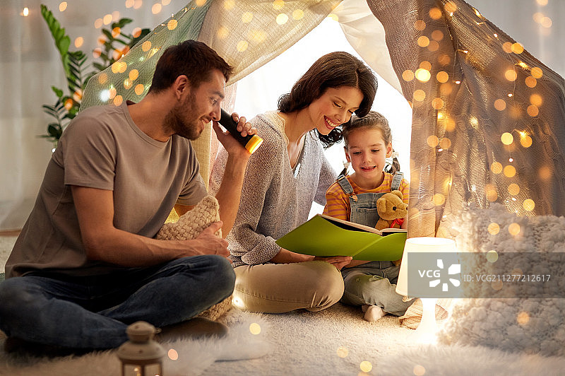 家庭、舒适和人的概念——快乐的妈妈、爸爸和小女儿晚上在家里的孩子帐篷里用手电筒看书。快乐的家庭在家里的孩子帐篷里看书图片素材