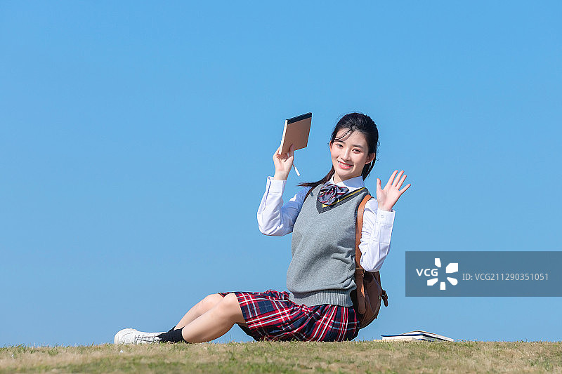 蓝色天空背景下，坐在草地上的亚洲女学生图片素材