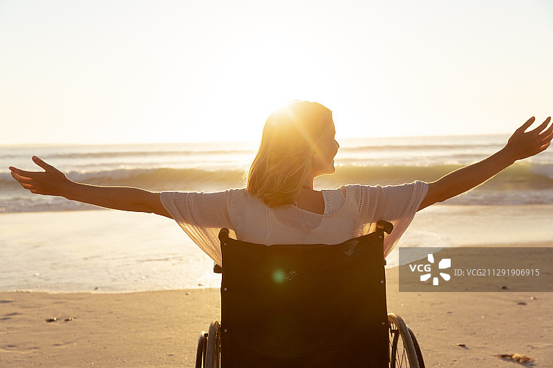 妇女在轮椅上的后视图与她的双臂在海滩上张开图片素材