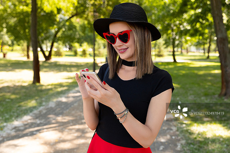 年轻女子戴着太阳镜和帽子，站在公园里使用手机图片素材