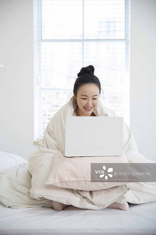 亚洲女人穿着睡衣在毯子下使用笔记本电脑图片素材