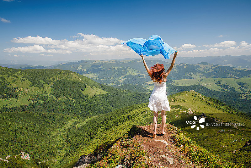 后视图的女人举起手臂与蓝色的围巾站在山上，对着天空图片素材