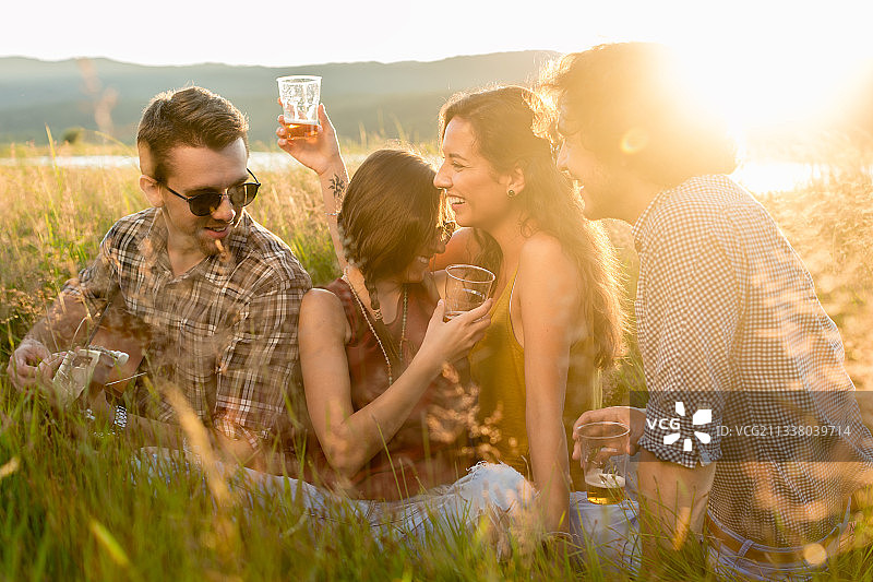 朋友们坐在高高的草地上，喝着饮料，听着吉他音乐，享受着夏日傍晚的心情图片素材