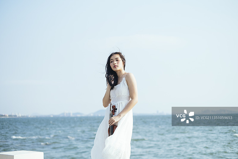 美丽的女人穿着白色的衣服站在海边，风吹着她的头发和衣服。图片素材