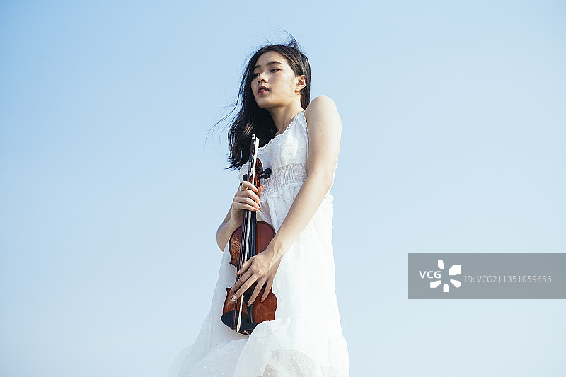 以天空为背景的黑色长发女子和她的小提琴的低角度拍摄。图片素材