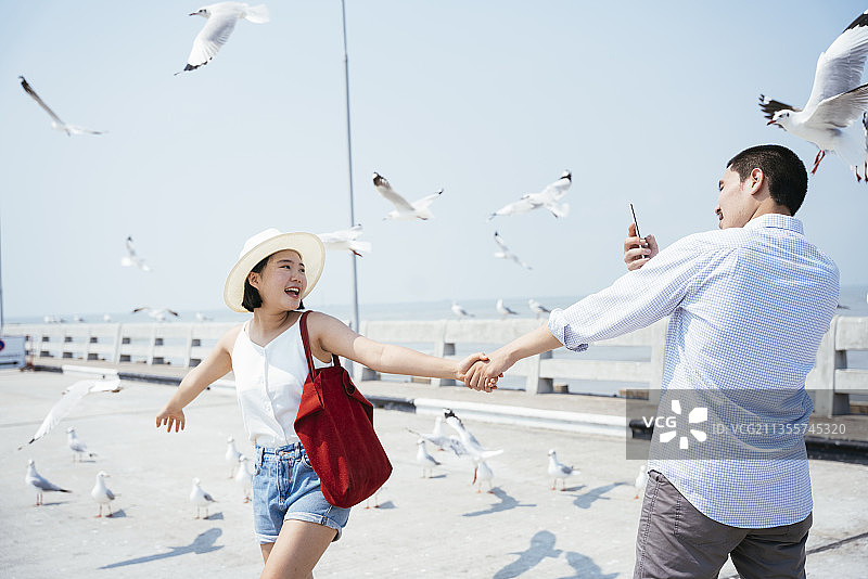 幸福浪漫的亚洲泰国情侣手牵着手，在一群海鸥中拿着智能手机自拍。图片素材