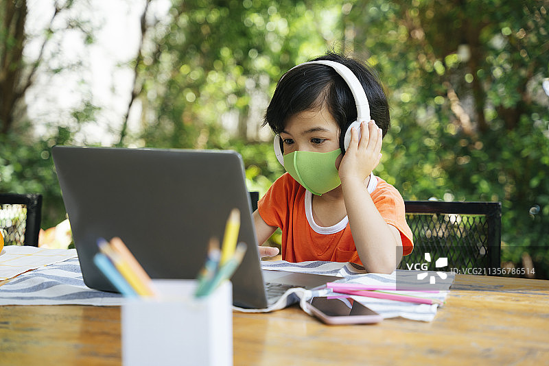 亚洲孩子戴着口罩，戴着耳机听音乐，在院子里使用笔记本电脑。图片素材