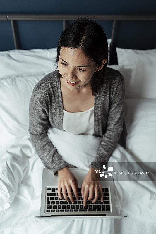 俯视图黑色短发的女人坐在白色的床上，带着笔记本电脑，微笑着在早上做她的工作。图片素材