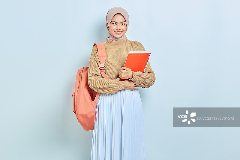 微笑的年轻亚洲穆斯林女学生在棕色毛衣和背包拿着书在白色的背景图片素材