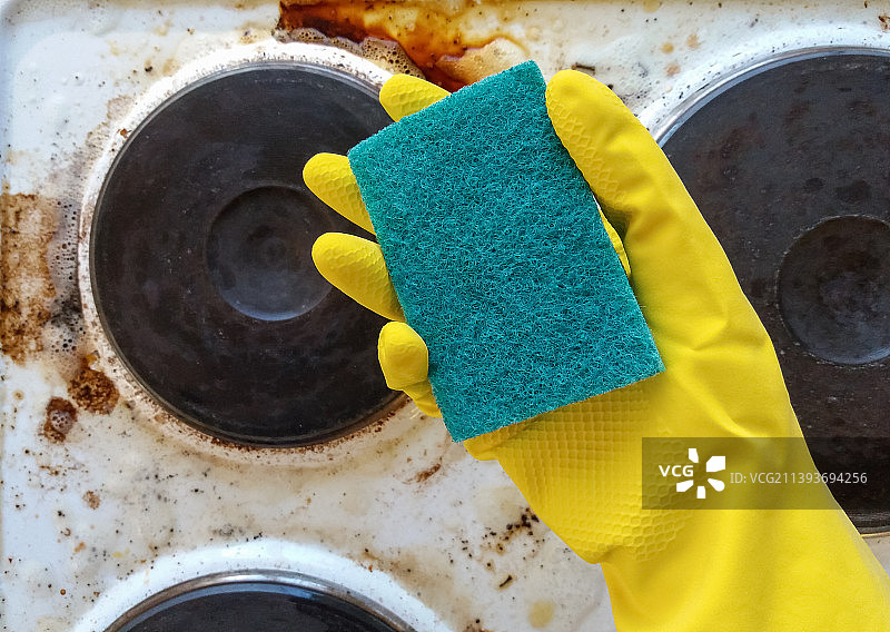 戴着黄色手套的手拿着洗碗海绵，对着脏兮兮的电炉图片素材