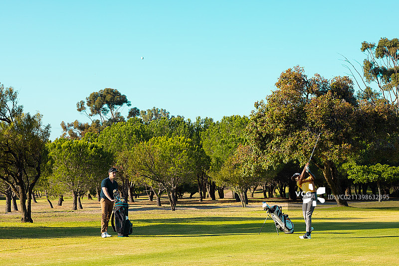 多种族的年轻男性朋友在夏天的高尔夫球场对着树木和晴朗的天空打高尔夫球图片素材