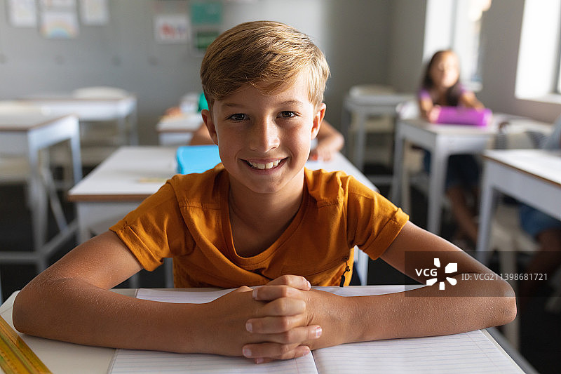 白人小学生微笑着双手合十坐在教室课桌前的肖像图片素材