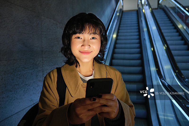 年轻的日本女孩拿着智能手机站在扶梯上图片素材