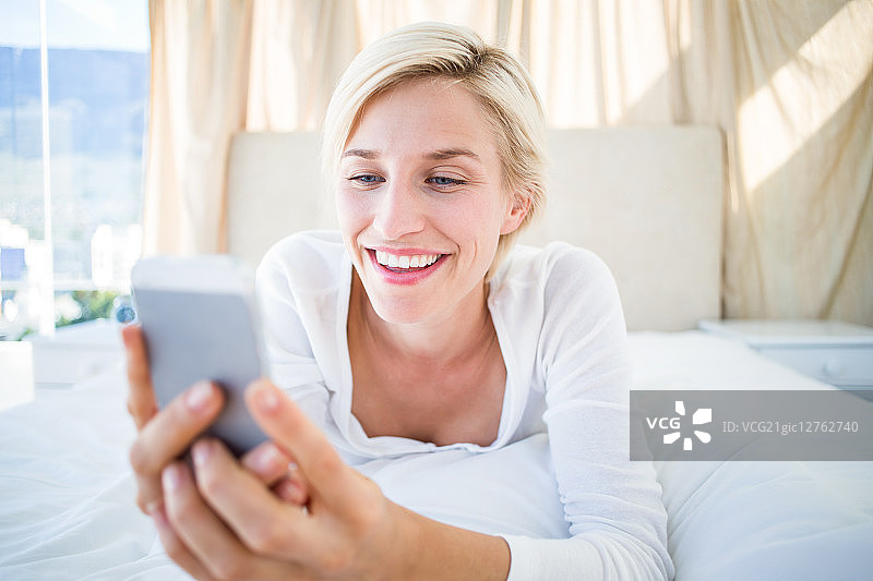微笑的金发女人躺在床上，用她的手机在卧室发短信图片素材