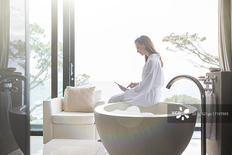 穿着白色浴衣的女人坐在现代浴缸边缘，使用着数码平板电脑图片素材