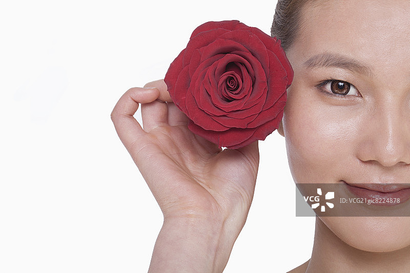 年轻女子微笑着，耳朵旁举着一朵红玫瑰，在摄影棚拍摄图片素材