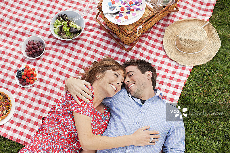 一对幸福的夫妇手挽手躺在野餐毯子上，旁边放着水果碗，就在正上方图片素材