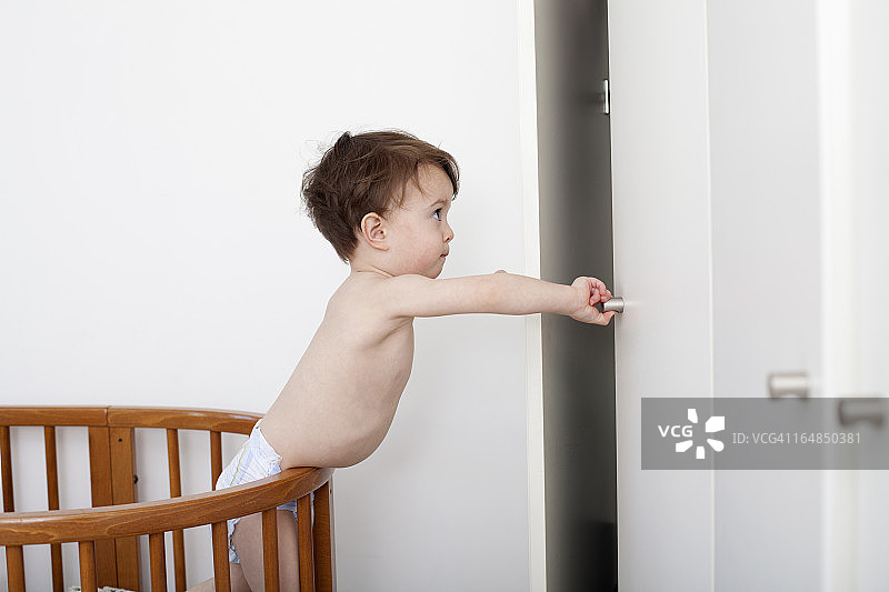 一个淘气的蹒跚学步的孩子从他的婴儿床上伸出手去打开壁橱的门图片素材