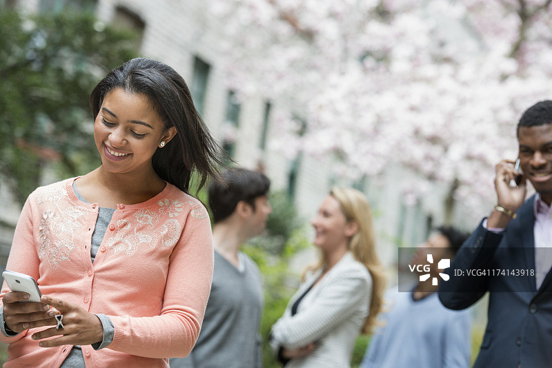 春天的城市生活。年轻人在城市公园的户外活动。一个穿粉红色衬衫的女人在看她的手机。背景中有四个人。图片素材