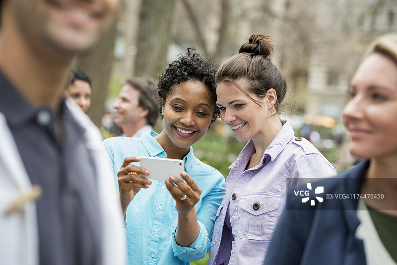 春天人们在城市里户外活动。纽约城市公园。两个女人在一群朋友，看着手机和微笑。图片素材