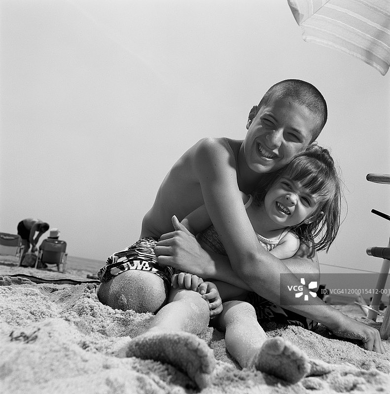 少年男孩(14-16岁)在海滩上拥抱妹妹(4-6岁)，肖像(B&W)图片素材