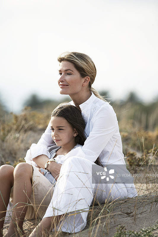 母亲和女儿(6-8)坐在长满草的沙丘上图片素材