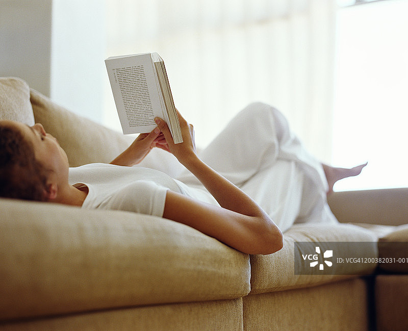 年轻女子躺在沙发上看书，低角度看图片素材