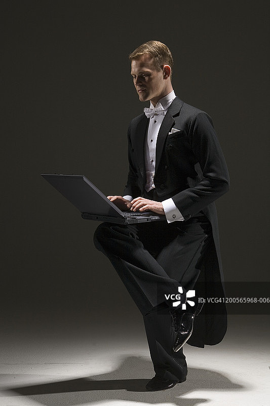 穿着燕尾服的男人一边用笔记本电脑工作一边用一只脚保持平衡图片素材