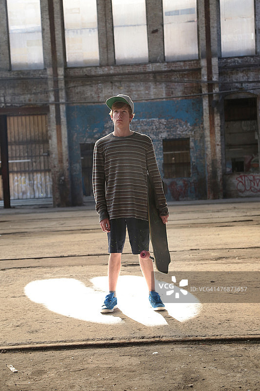 一个十几岁的男孩站在一个废弃的仓库滑板的肖像图片素材