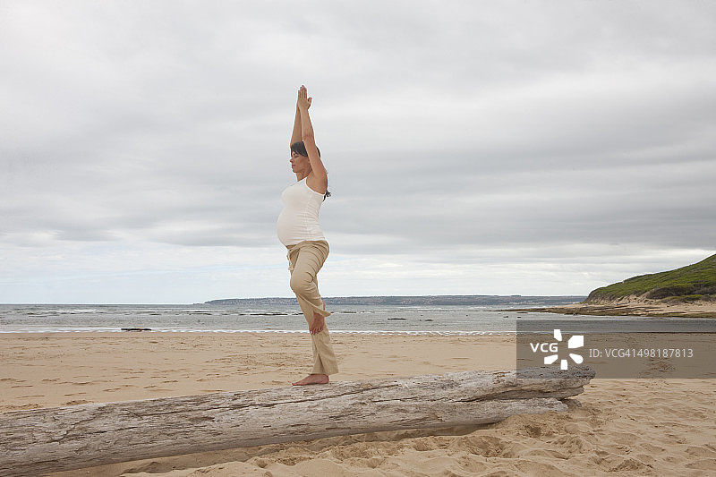 怀孕的中年妇女在海滩的树干上练习瑜伽图片素材