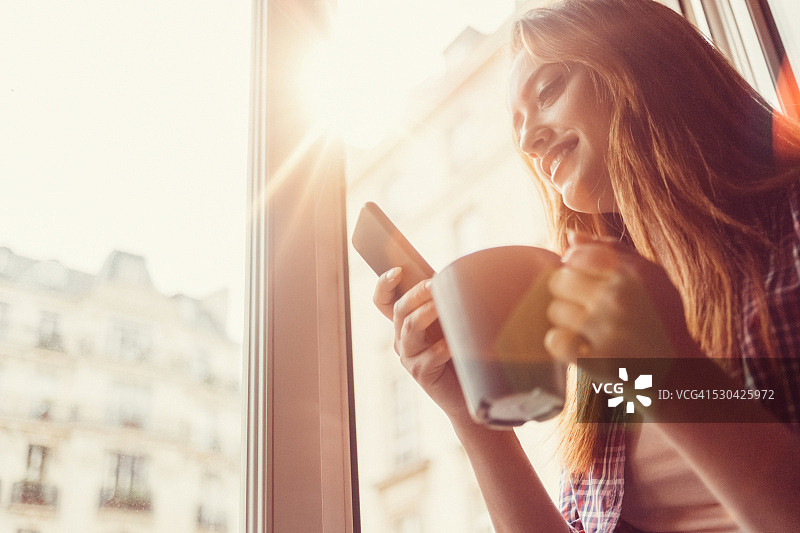 快乐的女人拿着咖啡杯在开着的窗户上发短信图片素材