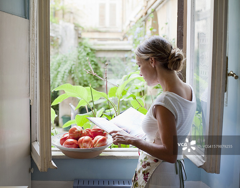 一个女人在打开的厨房窗户边看烹饪书图片素材