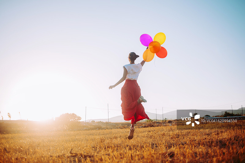 年轻女子拿着彩色气球在田野里快乐地跳跃图片素材
