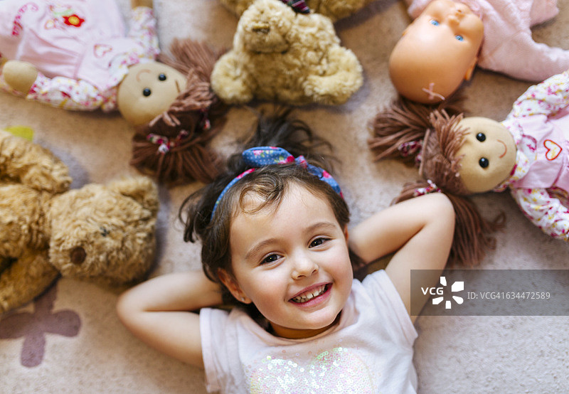一个微笑的小女孩的肖像躺在地板上，周围是泰迪熊和洋娃娃图片素材